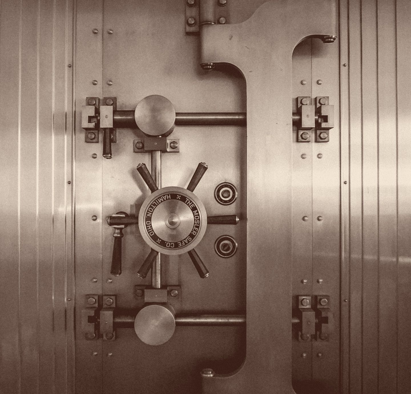 The closed steel door of a bank vault, photographed head-on: a spoked handwheel sits at the centre above a combination dial reading The Mosler Safe Co., Hamilton, Ohio, flanked by heavy locking bars and riveted hinges.