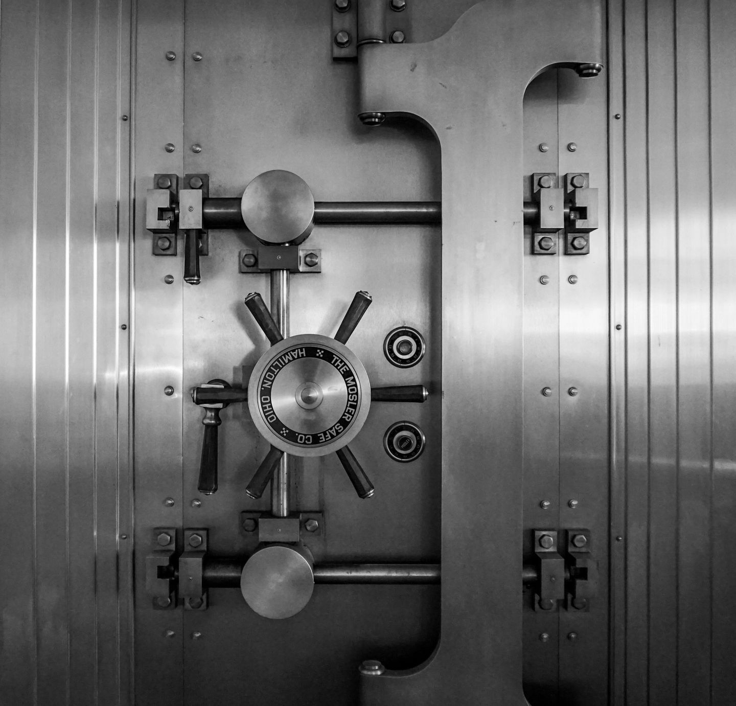 The closed steel door of a bank vault, photographed head-on in black and white: a spoked handwheel sits at the centre above a combination dial reading The Mosler Safe Co., Hamilton, Ohio, flanked by heavy locking bars and riveted hinges.